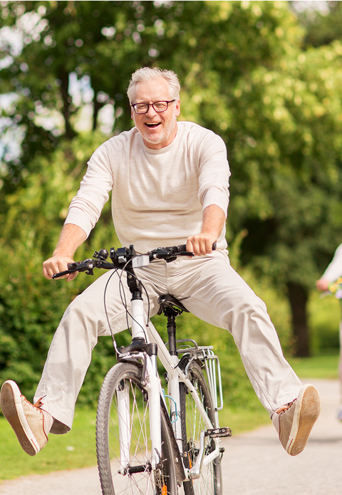 Older man riding a bicycle outdoors with his legs stretched out and smiling, wearing glasses and casual light-colored clothes. Green trees and path in the background.