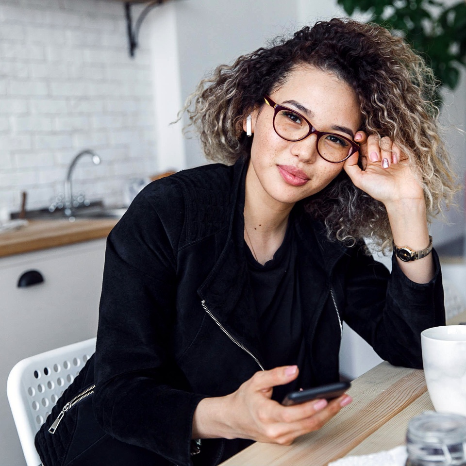 curly-haired girl in a black jacket in an apartment at home in the studio stands in the kitchen near the table with a cup in her hands posing and smiling laughter