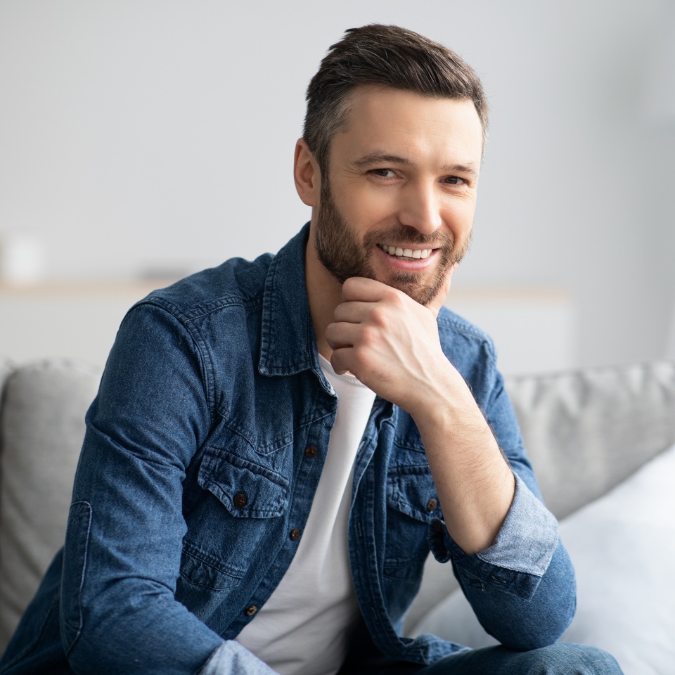 Closeup portrait of handsome bearded man sitting on couch and smiling, home interior. Cheerful middle-aged man relaxing in living room, touching his ching and smiling at camera, copy space