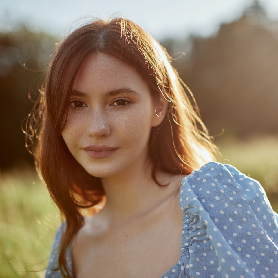 Close up of a beauty young woman in the sun rays. Redhead girl looking at the camera wearing blue dress.