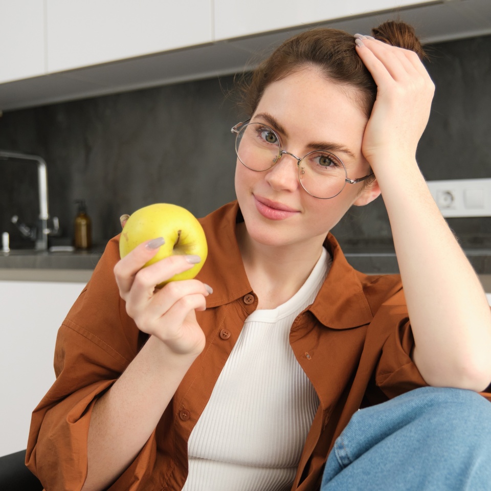 Portrait of smiling, modern woman in glasses, sitting at home and eating green apple, having a lunch break, biting fruit.