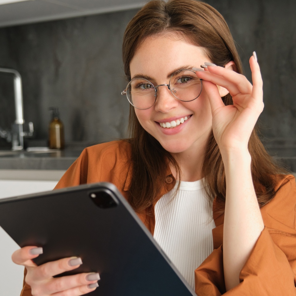 Portrait of confident, smiling young woman, student in glasses, sitting in kitchen, studying on remote, connects to online course on digital tablet.