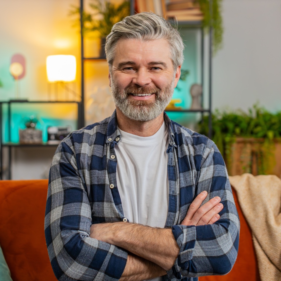 Happy Caucasian senior man sitting on sofa looking at camera and smiling at home. Bearded mature guy having attractive appearance enjoying carefree retired life relaxing in living room in apartment.