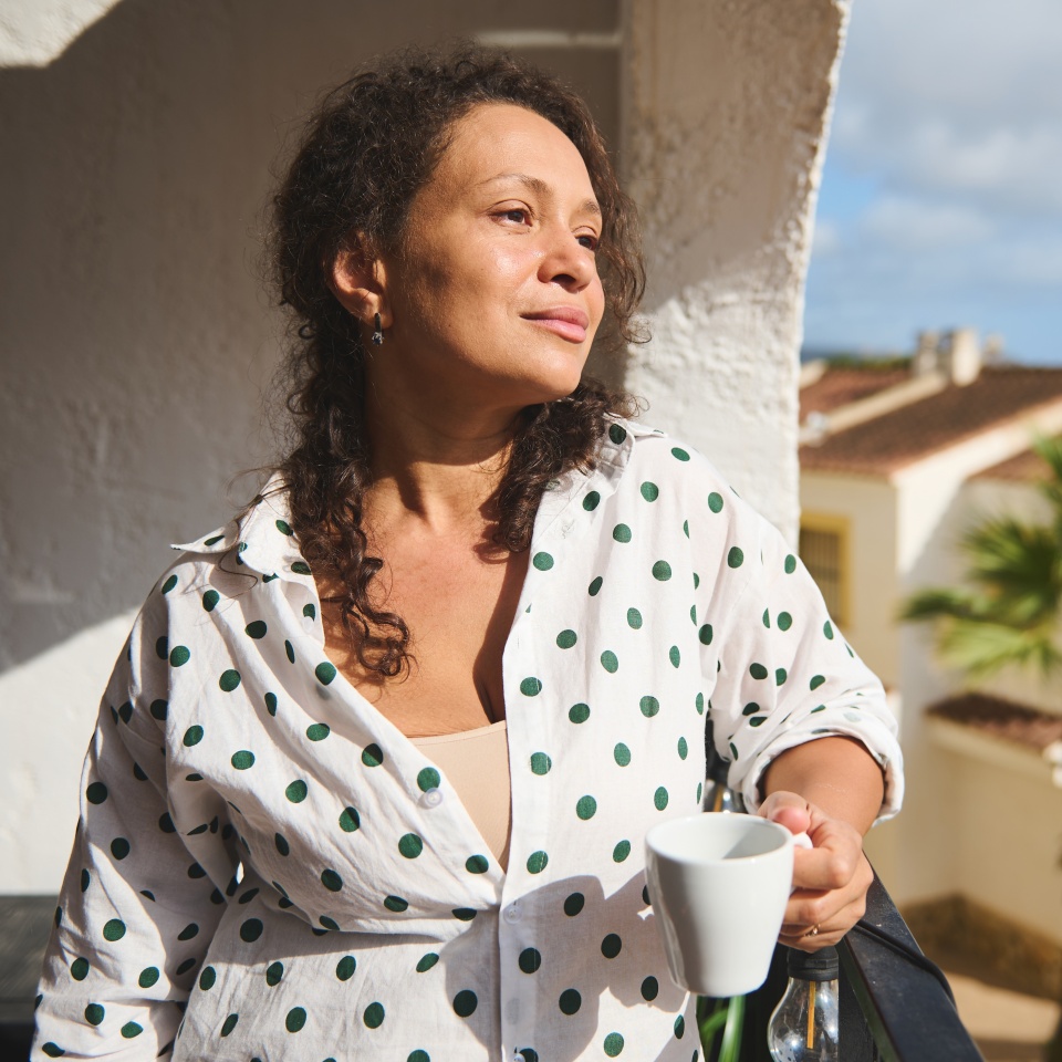 A woman stands on a sunny balcony, savoring a warm cup of coffee. She overlooks picturesque Mediterranean-style homes and lush palm trees, enjoying a peaceful morning moment.