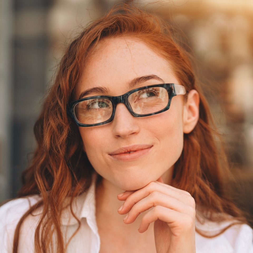Young woman with red hair wearing glasses ponders creatively at an outdoor cafe in the afternoon light