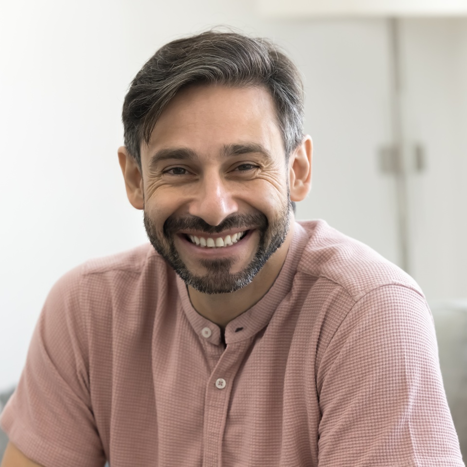 Handsome middle-aged man with warm smile, wear light pink shirt posing for camera, head shot portrait view. Portuguese male looks relaxed, contented, resting alone seated on cozy sofa in living room