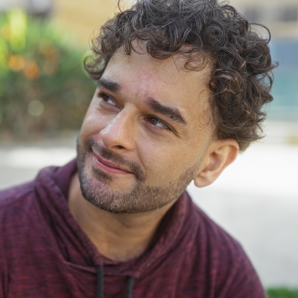 Handsome man with curly hair smiling in an urban park, surrounded by greenery, capturing a serene moment in a bustling city environment, sunlight gently illuminating his face.