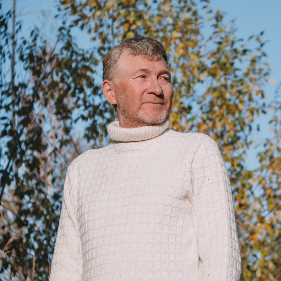 handsome man middle aged in cozy white sweater enjoying crisp autumn day outdoors. surrounding him are vibrant autumn trees with golden and red leaves under clear blue sky. close up.