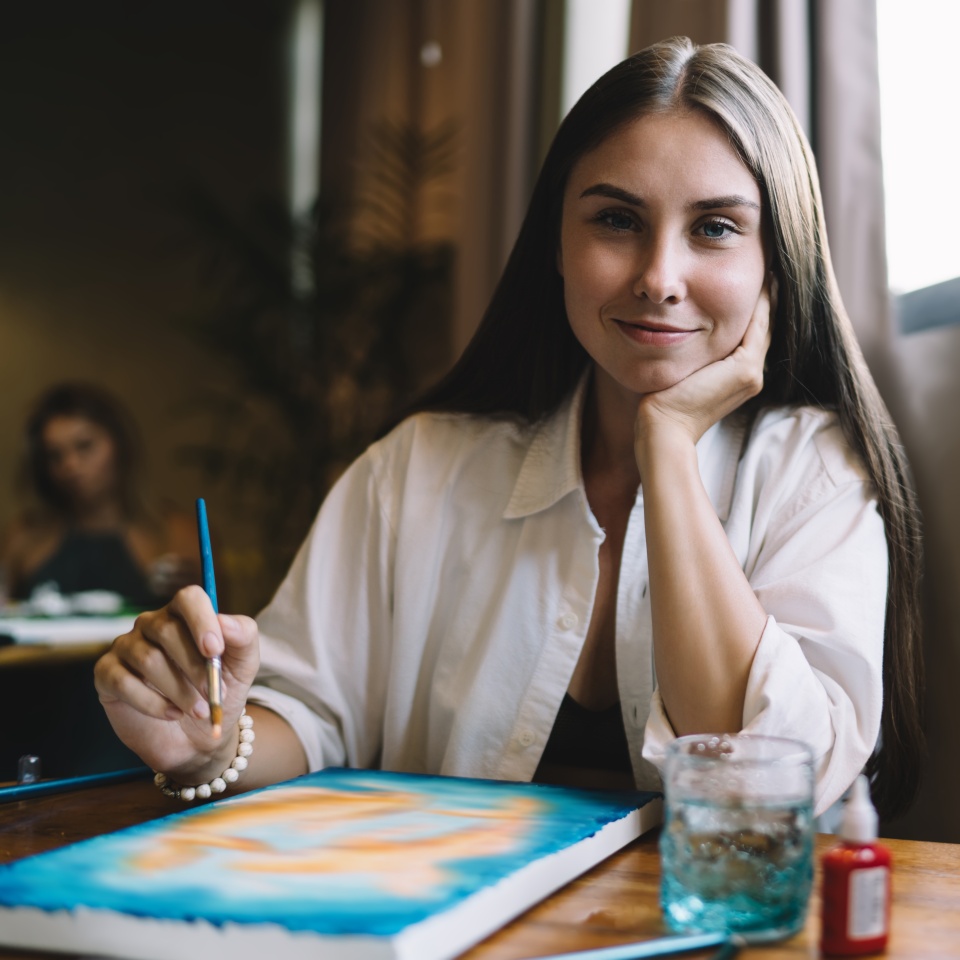 Calm woman rests her face on hand while holding brush, smiling softly—symbolizing quiet joy, self-connection, artistic identity and tech-enabled personal growth.