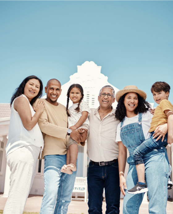 A group of six people, including adults and children, stand smiling outdoors in front of a white building under a clear blue sky.