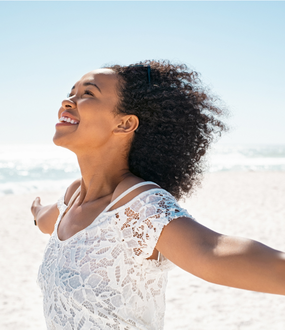 Woman in a white lace top stands on a sunny beach with arms outstretched, smiling and looking upwards.