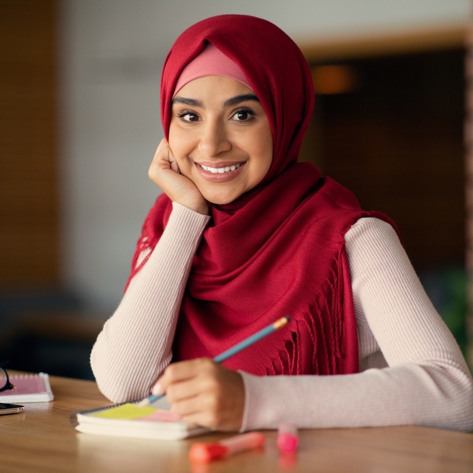 Cheerful pretty young muslim businesswoman in hijab planning day, writing notes while drinking coffe alone at cafe, leaning on her hand and smiling at camera, copy space, closeup portrait