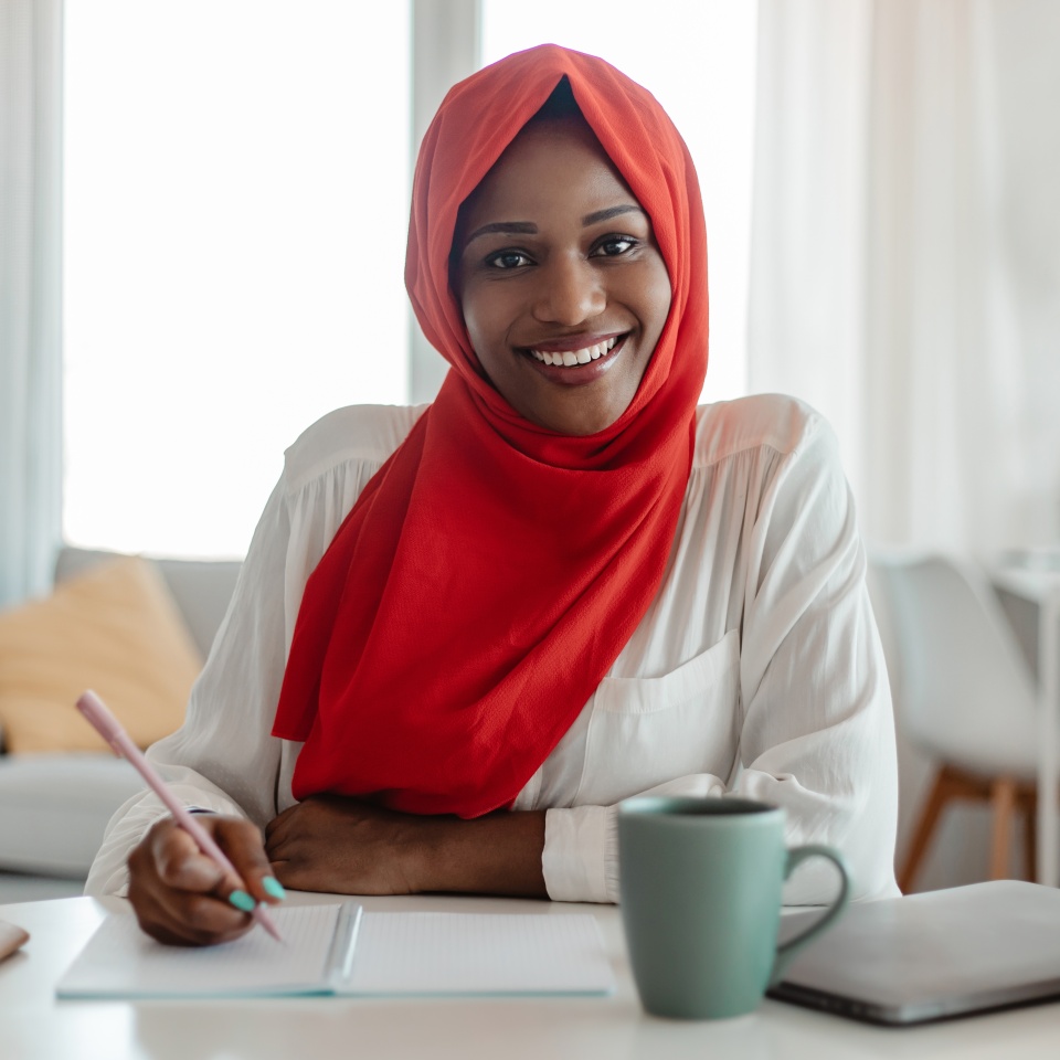 Positive black muslim woman independent contractor sitting at workdesk with laptop and cup of coffee at home office, writing at notepad and smiling. Lady planning her working day