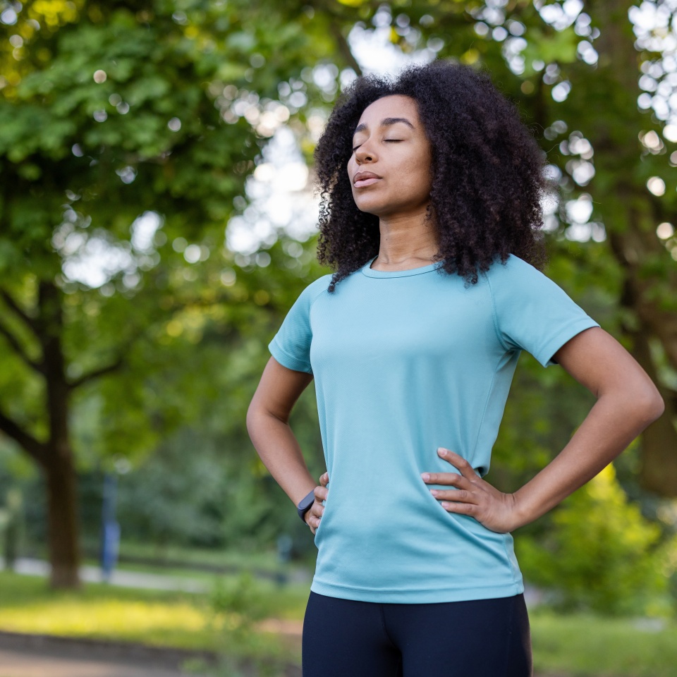 African American female practicing mindfulness and relaxation in park. Standing confidently with eyes closed and hands on hips. Emphasizing fitness, wellbeing, mental clarity, outdoor lifestyle