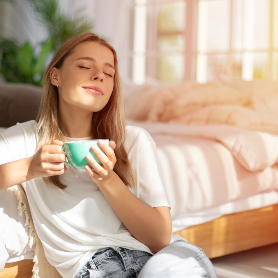 Young woman relaxes in sunlight, holding teal mug with eyes closed, enjoying a peaceful moment in a cozy bedroom. The room is bathed in natural light, featuring soft bedding, evoking calm and warmth