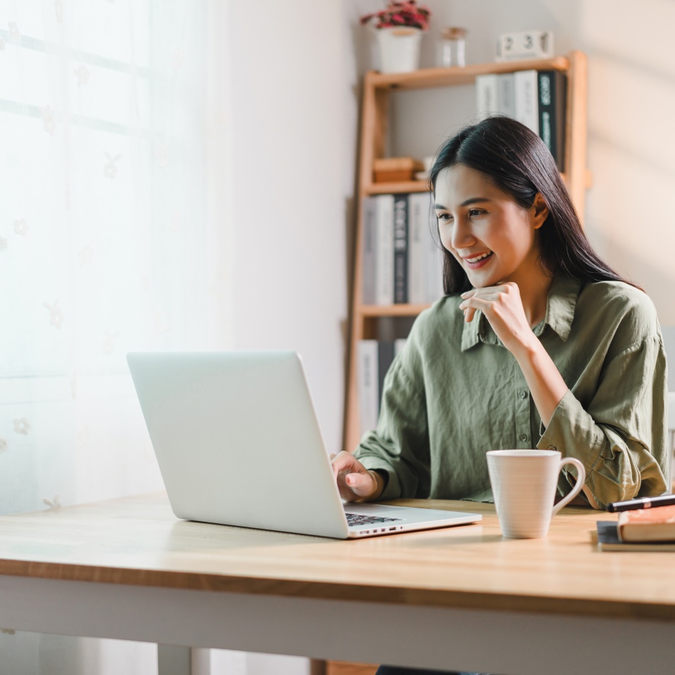 A woman working on laptop at wooden desk in bright home office, smiling while holding cup of coffee. room features bookshelves, lamp, and natural light from window