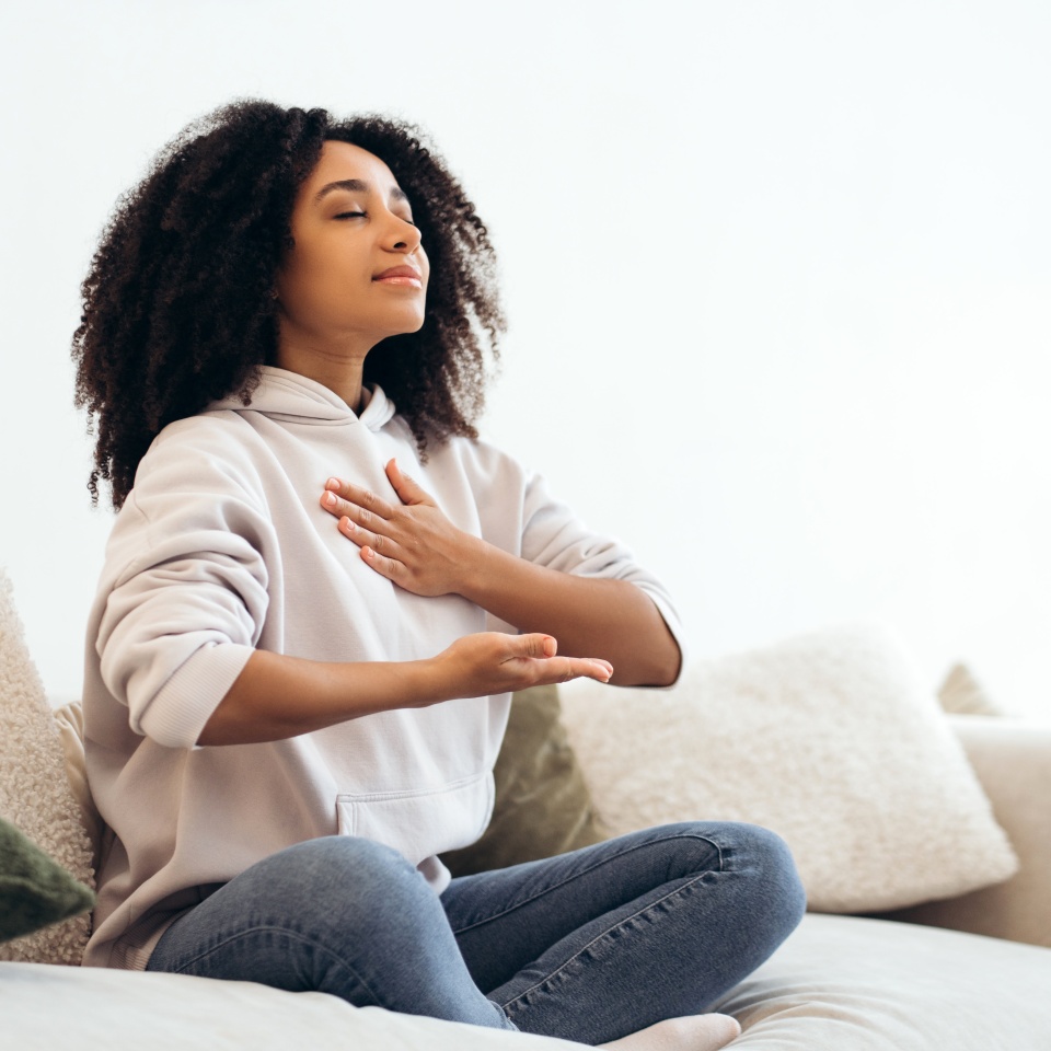 Serene young woman practicing breathing exercises while sitting in lotus position on comfortable sofa at home, enjoying meditation and relaxation