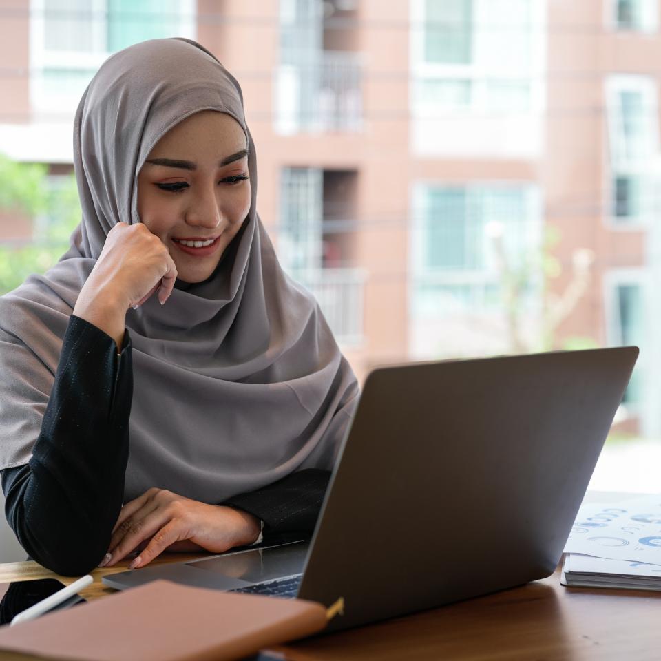 A woman wearing a hijab sits at a desk, smiling while looking at a laptop. There are books, papers, and a pen on the desk. A window with a building outside is in the background.