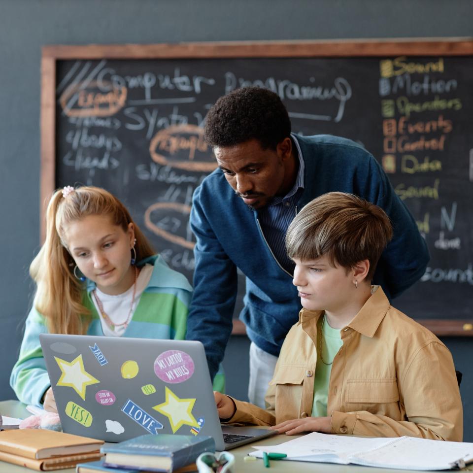 A teacher helps two students working on a laptop in a classroom, with a chalkboard displaying computer programming notes in the background.