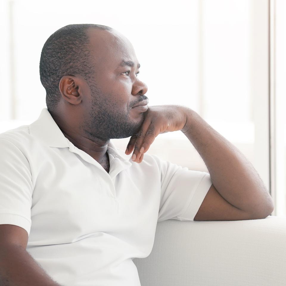 A man in a white polo shirt sits on a couch, resting his chin on his hand, looking thoughtfully out of a bright window.