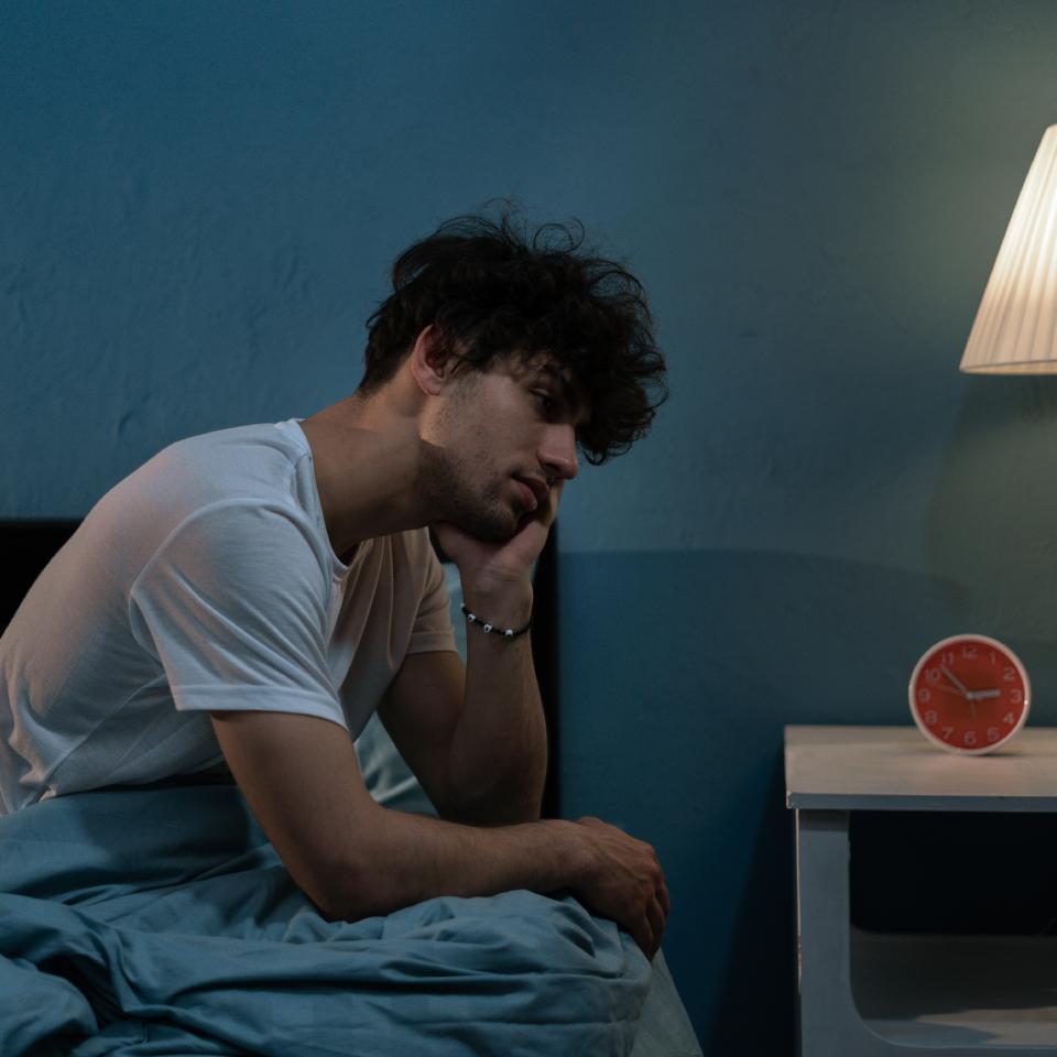 A man in a white t-shirt sits on a bed, looking tired, with one hand on his face. A lit lamp and a red alarm clock are on the bedside table.