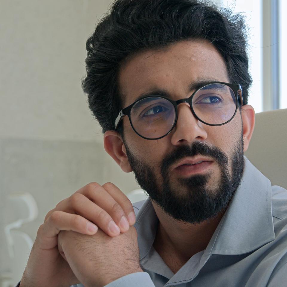 A man with dark hair, a beard, and glasses sits indoors, clasping his hands and looking thoughtfully to the side. He is wearing a light blue shirt.