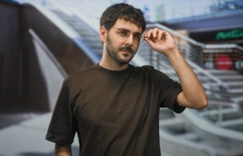 Man adjusts glasses with right hand in airport terminal by escalator and railing, looking thoughtful and pensive; calm contemplation.